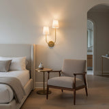 Greta Double Wall Sconce, Satin Brass - A satin brass double wall sconce illuminates a bedroom with a bed, chair, and side table.