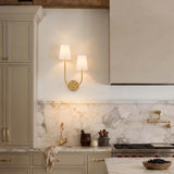 A transitional kitchen featuring a Greta Double Wall Sconce, Satin Brass with linen shades, mounted on a marble backsplash beside taupe cabinets under a beamed ceiling.