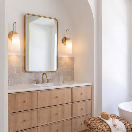 A transitional bathroom with two Connie Wall Sconce 17", Rustic Brass with Opal Glass fixtures flanking a gold-rimmed mirror above a light wood vanity and tiled backsplash.