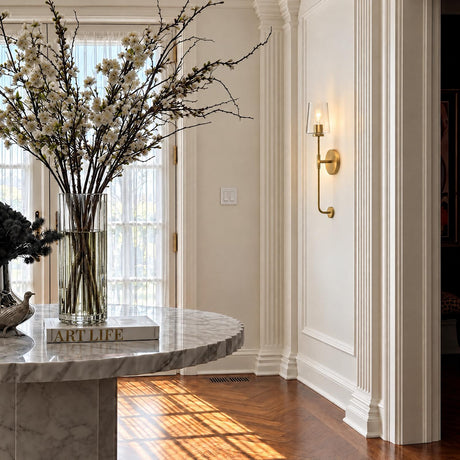 A transitional foyer featuring a Wyatt Wall Sconce 23", Satin Brass with Clear Glass mounted on a white paneled wall beside herringbone wood floors and marble table.
