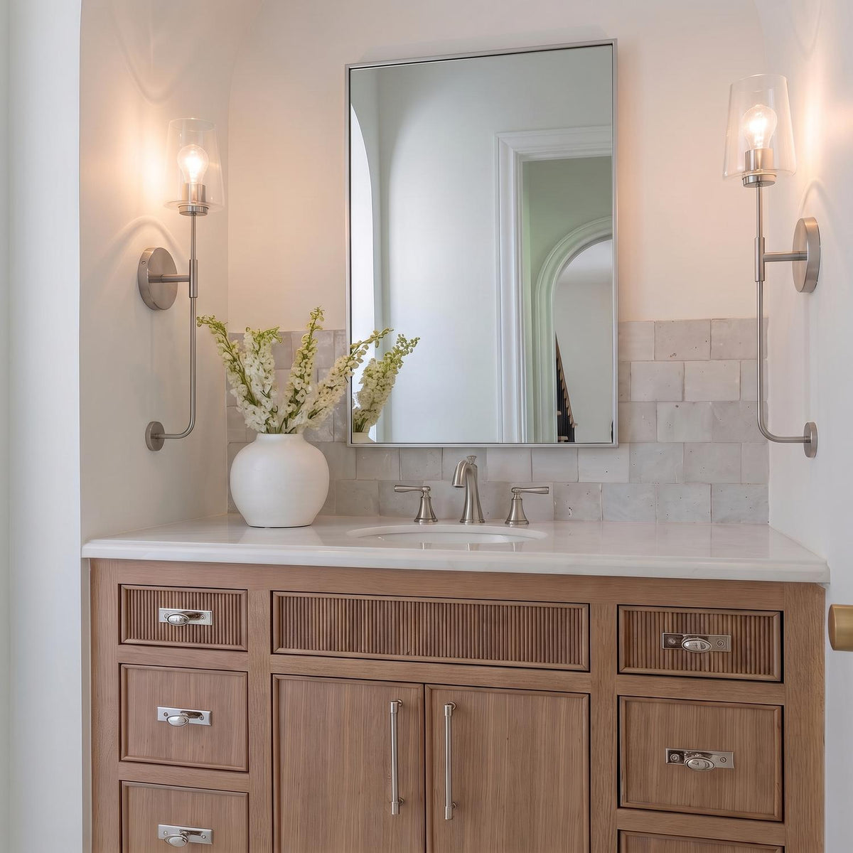 A transitional bathroom with two Wyatt Wall Sconce 23", Brushed Nickel with Clear Glass fixtures flanking a mirror over a light wood vanity and white marble countertop.