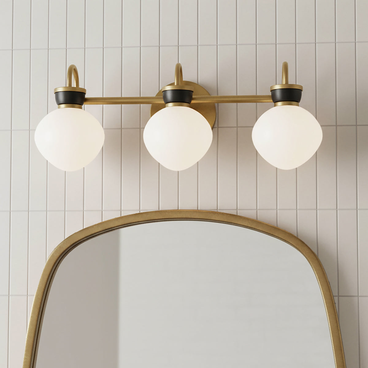 Piper 3 Light Vanity, Satin Brass and Black - A three-light vanity fixture with globe shades is mounted above a gold-framed mirror against a white tiled wall.