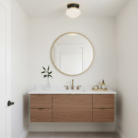 Piper Flush Mount 12, Satin Brass and Black - A flush mount light fixture with a black base and frosted glass shade is mounted on a white ceiling above a round gold mirror and a floating wood vanity in a bathroom.