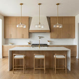A modern kitchen features three Alder Pendant 15", Satin Brass with Clear Glass fixtures suspended over a white marble waterfall island and light wood cabinets.