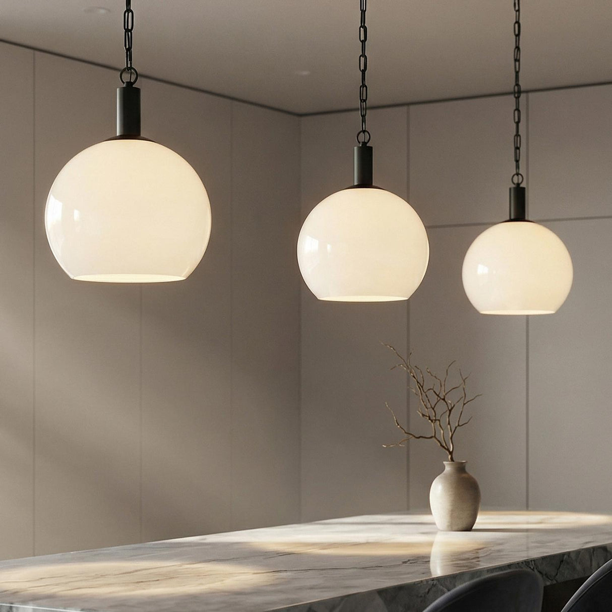 A minimalist kitchen features three Jolie Pendant 13", Black fixtures with matte black hardware and white glass globes over a white marble island with beige cabinetry.