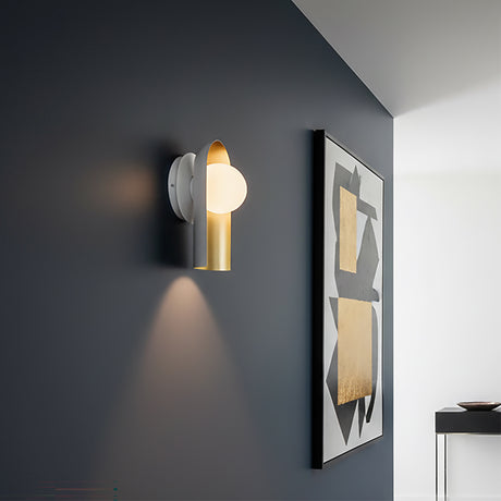Kassidy Wall Sconce 10 - White and Satin Brass wall sconce with a modern design against a dark gray wall, illuminated by a warm glow.