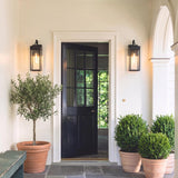 A modern farmhouse porch with two matte black Monterey Outdoor Wall Light lanterns glowing beside a dark front door, flanked by terracotta potted plants and white siding.