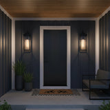 Monterey Outdoor Wall Light, Large 25" Black - Two black lanterns flanking a dark gray door on a covered porch with wooden ceiling, a bench, plants, and a doormat.