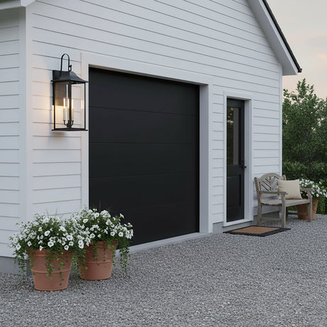 Lorraine Outdoor Wall Light, Large 26" Black - A black lantern-style outdoor wall light illuminates the white siding of a garage with a black door, flanked by potted white flowers.