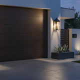Jameson Outdoor Wall Light - A black wall lantern with brass accents illuminates the exterior of a modern home next to a dark wood garage door.