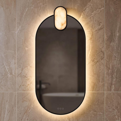 A contemporary bathroom with the Palmer Large LED Oval Mirror in matte black and alabaster mounted to tan marble wall tiles reflecting a white tub.