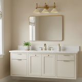 Fletcher 3 Light Vanity, Patina Brass and Soft Sand - A vanity light with three conical shades in a soft sand color hangs above a large rectangular mirror with a brass frame, situated above a cream-colored vanity with brass hardware and a white countertop.