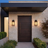 A modern entry with two Durham Outdoor Wall Light lanterns in a textured black finish flanking a dark wood door on white stone at twilight with boxwood bushes.