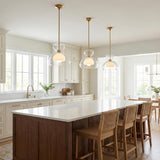 A transitional kitchen features three Rhett Pendant 12", Aged Brass fixtures with clear and frosted glass suspended over a white quartz island with woven stools.