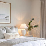 A coastal bedroom featuring the Calista Ceramic Table Lamp, Whitewashed Terracotta with a white linen shade on a nightstand beside textured pillows and framed beach photography.