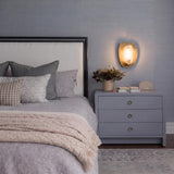 A contemporary bedroom featuring the Jane Wall Sconce 12", Patina Brass with a frosted glass center mounted on a textured blue wall above a grey nightstand.
