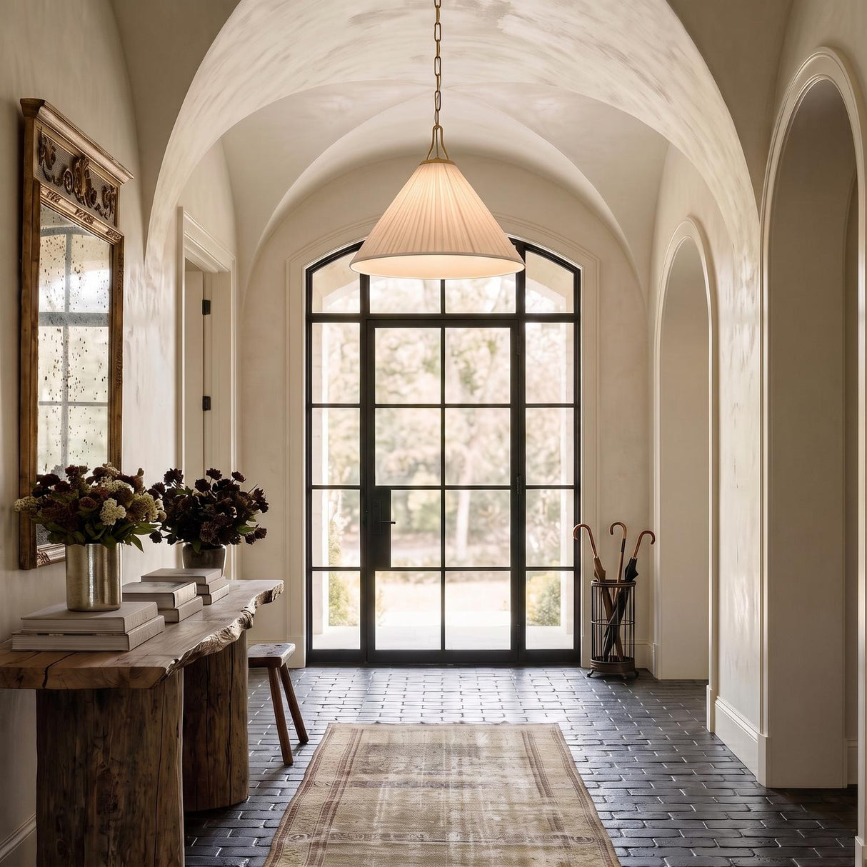 A Mediterranean foyer with the Charlotte Medium Pendant 22", Aged Brass featuring a pleated linen shade hanging from a vaulted ceiling above dark brick flooring.