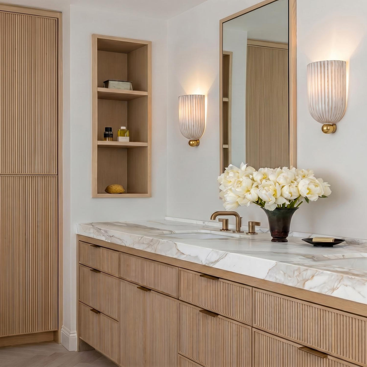 A modern bathroom with two Meredith Wall Sconce 10", Aged Brass and Ceramic fixtures flanking a mirror above a marble countertop and slatted oak vanity.