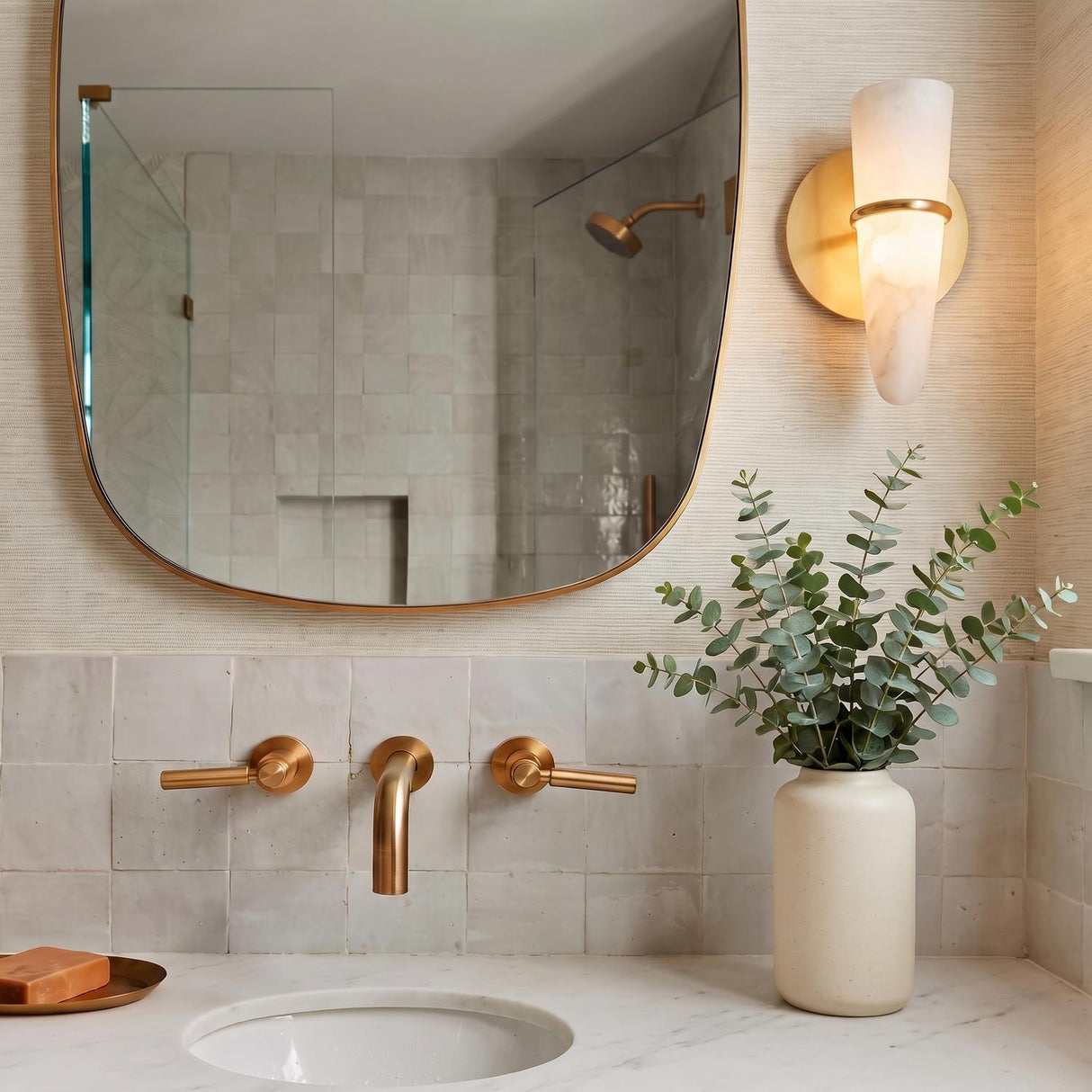 A transitional bathroom with the Fira LED Wall Sconce 10", Vintage Brass and Alabaster, mounted beside an arched mirror above a marble vanity and brass faucet.
