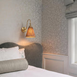 A traditional bedroom featuring an aged brass Rhiannon Wall Sconce with a pleated rattan shade mounted beside a grey velvet headboard and floral wallpaper.