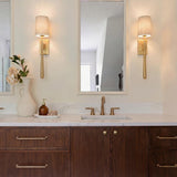 A transitional bathroom featuring two Norene Wall Sconce 25", Aged Brass fixtures with linen shades flanking a mirror above a marble vanity and walnut wood cabinets.