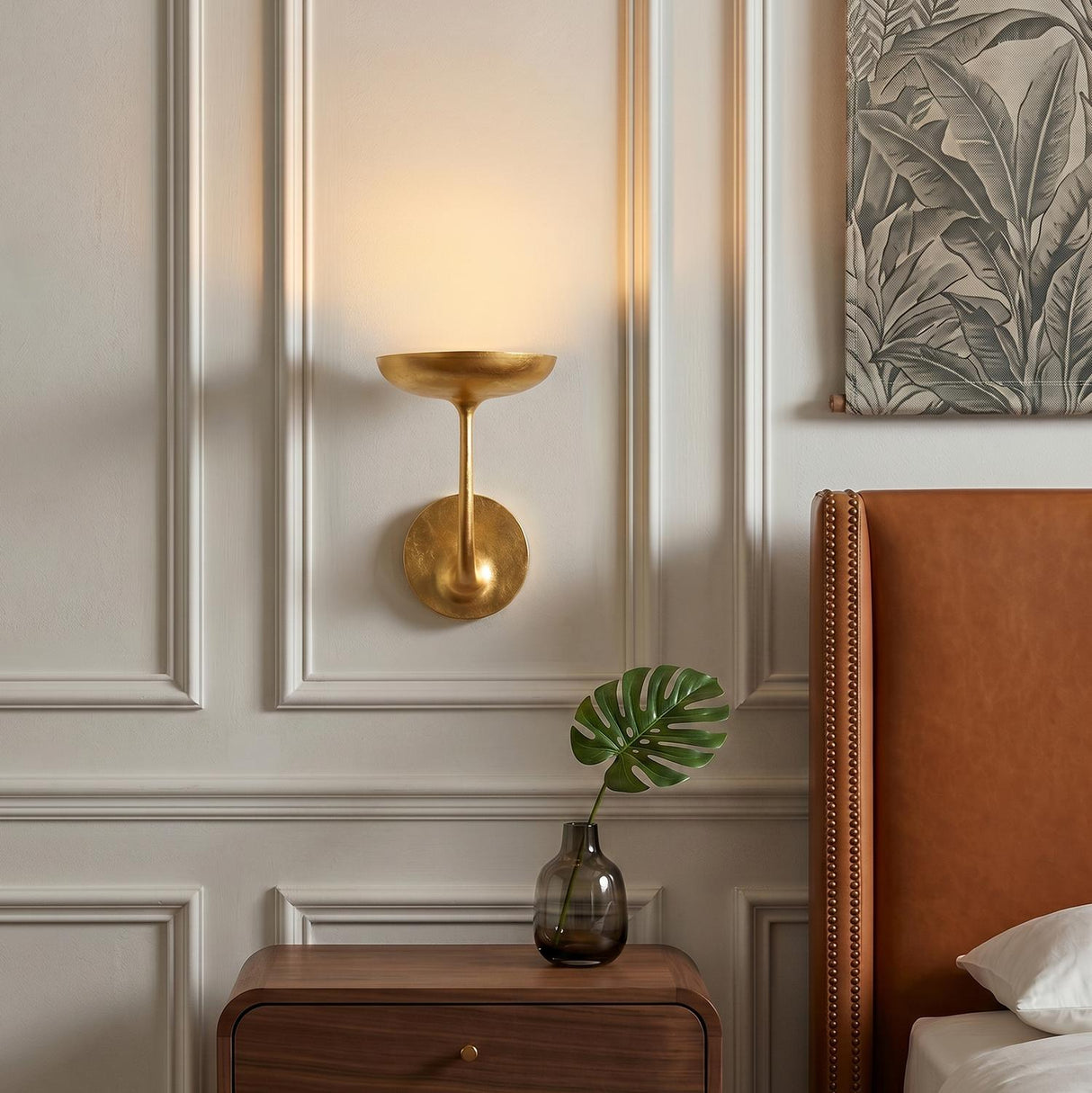 A transitional bedroom with a gold leaf Reverie LED Wall Light 9" on a cream paneled wall beside a cognac leather headboard and walnut nightstand.