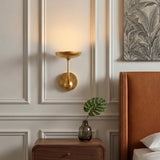 A transitional bedroom with a gold leaf Reverie LED Wall Light 9" on a cream paneled wall beside a cognac leather headboard and walnut nightstand.