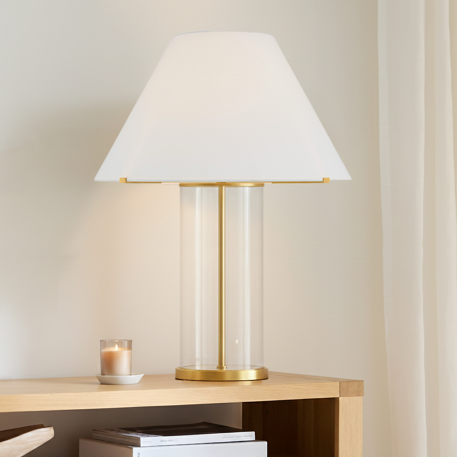 Table lamp is 30" tall with white shade and clear glass with aged brass base, on a wooden console table with books and a candle.