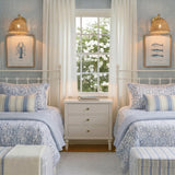 A coastal bedroom with two Karina Wall Sconce 8", Woven Wicker and Aged Brass fixtures mounted above twin beds with blue floral bedding and a white nightstand.