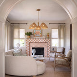 A transitional living room features a Karina 5 Light Chandelier in woven wicker and aged brass suspended above a pink checkered tile fireplace and curved cream sofa.