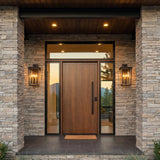 A modern home entry with Radcliffe Outdoor Wall Light lanterns in old bronze flanking a wood door on a stacked stone facade with boxwood at twilight.