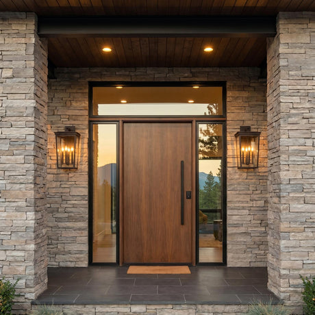 A modern home entry with Radcliffe Outdoor Wall Light lanterns in old bronze flanking a wood door on a stacked stone facade with boxwood at twilight.