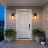 A modern farmhouse porch featuring two old bronze Radcliffe Outdoor Wall Lights flanking a white front door on tan siding with potted ferns and flowers at dusk.