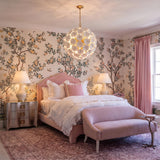 A traditional bedroom with a white ceramic floral chandelier in a gold finish hanging from the ceiling center above a pink upholstered bed and botanical wallpaper.