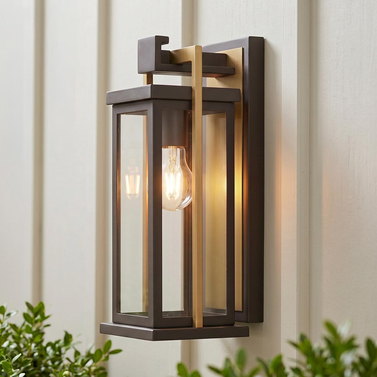 A modern farmhouse patio featuring the Quinlan Outdoor Wall Light, Small 15", Bronze and Textured Gold glowing at dusk on white board-and-batten siding above green boxwood shrubs.