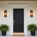 A colonial home entry with bronze and textured gold Quinlan Outdoor Wall Light lanterns flanking a black front door on white brick at dusk with potted ferns.