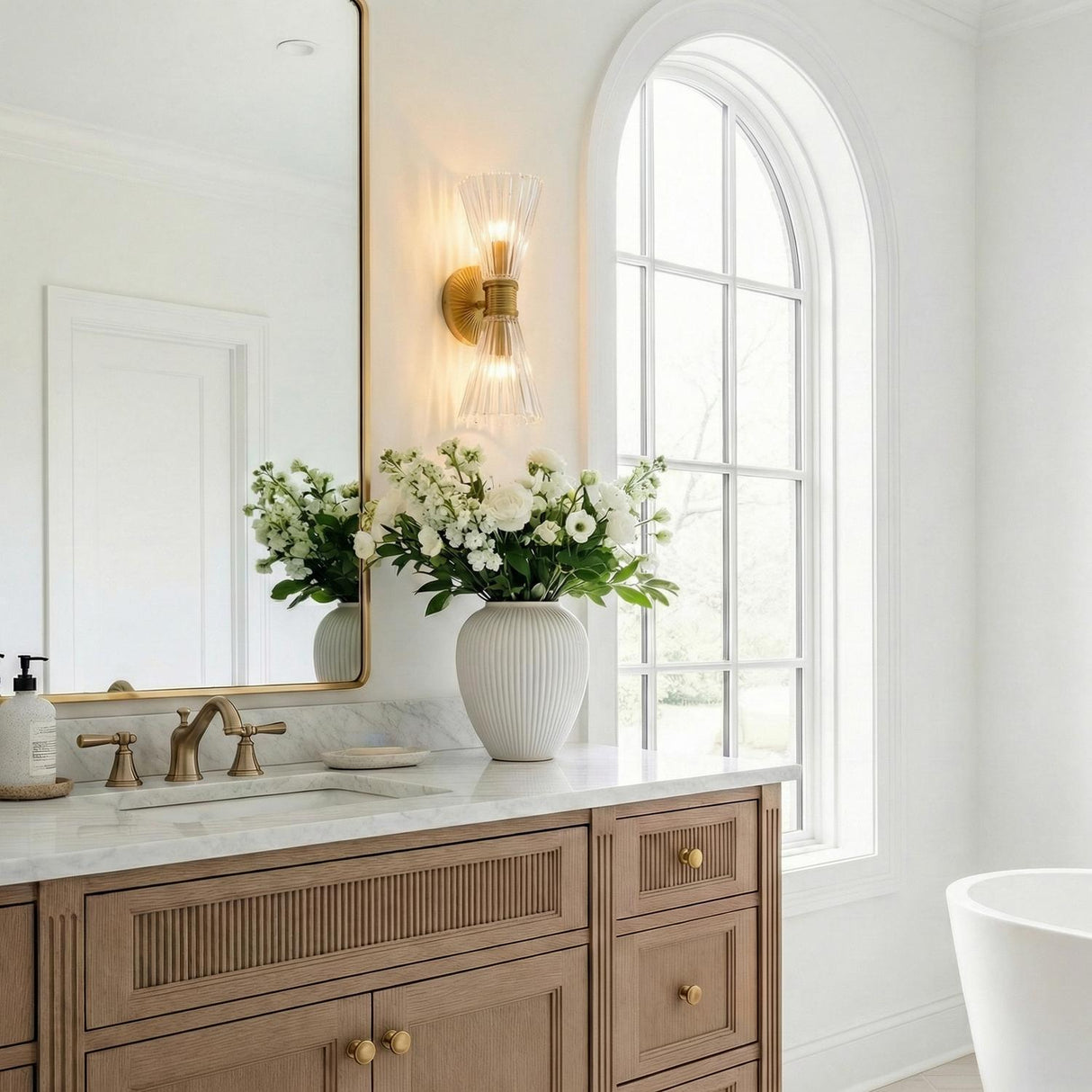 Eliza 2 Light Vanity, Brass and Clear Ribbed Glass ribbed Wall Sconce in a Contemporary Bathroom with a wood vanity and arched window.