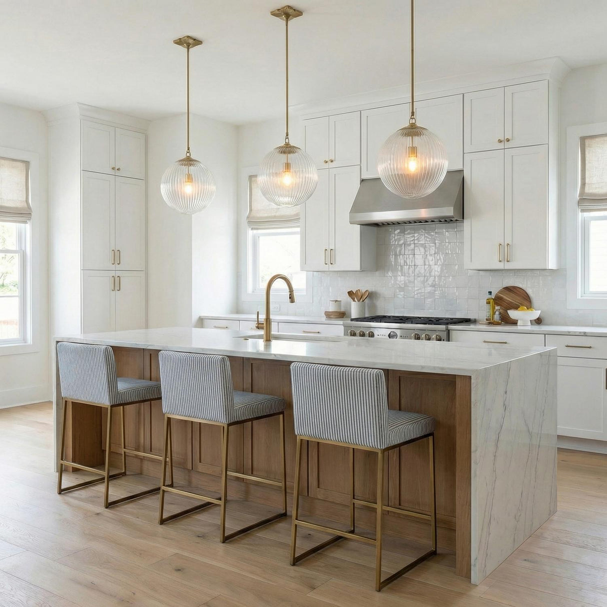 A transitional kitchen features three Victoria Pendant 14" in Aged Brass and Clear Ribbed Glass suspended over a white marble waterfall island with wood accents.