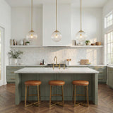 A transitional kitchen features three Victoria Pendant 14" in aged brass and clear ribbed glass over a white marble island with sage green cabinets and leather stools.