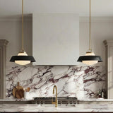 A contemporary kitchen features two Blaine Medium Pendant 16", Brass and Matte Black fixtures flanking a white range hood above a purple-veined marble backsplash.