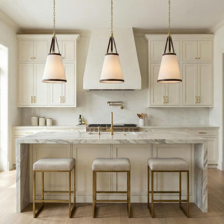 A transitional kitchen features three Spencer Small Pendant 14", Brass with Brown Leather Accents over a white marble waterfall island with white cabinets and linen shades.