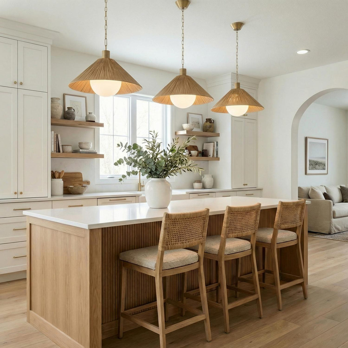 A coastal kitchen features three Fawn Medium Pendant 22 inch, Patina Brass and Rattan with Opal Glass fixtures over a fluted wood island with three woven bar stools.