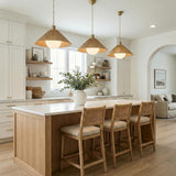 A coastal kitchen features three Fawn Medium Pendant 22 inch, Patina Brass and Rattan with Opal Glass fixtures over a fluted wood island with three woven bar stools.