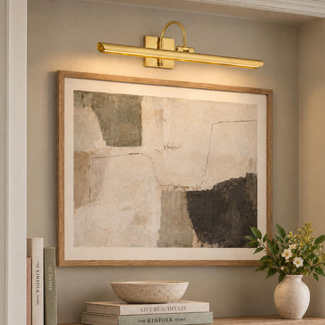 A transitional living room features the Carlin LED Picture Light 24", Aged Brass mounted above framed abstract artwork on a neutral wall above stacked books and flowers.