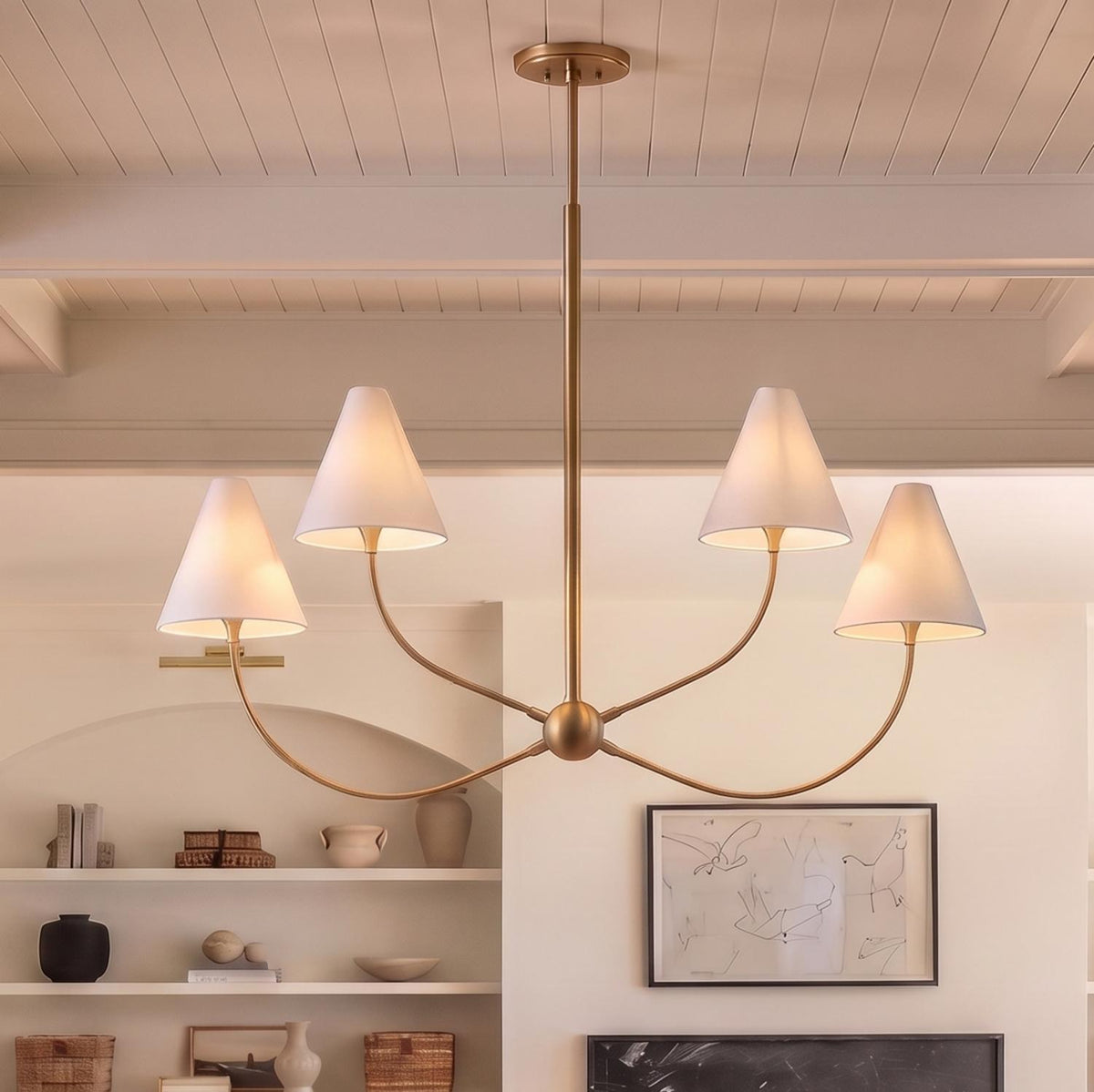 A transitional living room with a patina brass chandelier featuring four white linen shades suspended from a beamed ceiling over an arched shelving niche with neutral pottery.