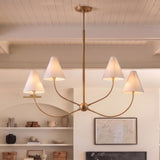 A transitional living room with a patina brass chandelier featuring four white linen shades suspended from a beamed ceiling over an arched shelving niche with neutral pottery.
