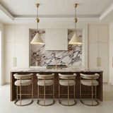 A contemporary kitchen features two Hadley Pull-Down Pendant 18", Soft Sand and Patina Brass fixtures over a marble island with wood fluting and four beige barstools.