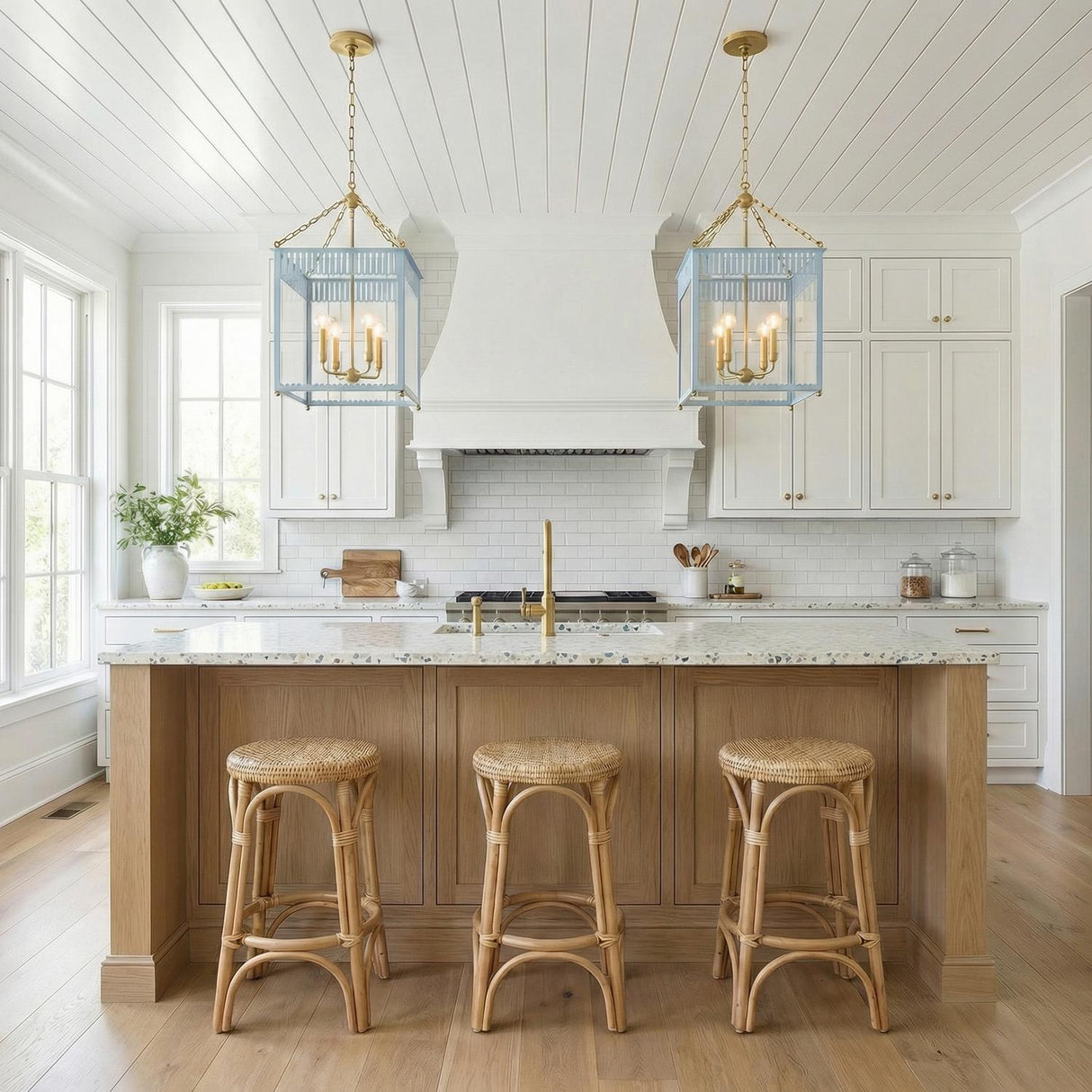 A coastal kitchen features two Roanne Large Lantern Pendant 17", Light Blue and Aged Brass fixtures hanging over a terrazzo island under a white shiplap ceiling.