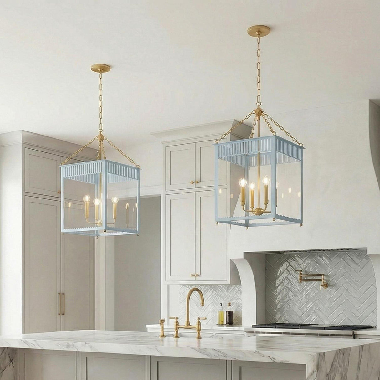 A transitional kitchen features two light blue and aged brass lantern pendants suspended over a white marble island with a herringbone tile backsplash and brass pot filler.