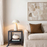 A contemporary living room featuring the Janna Table Lamp 21", Artisan Ivory on a dark wood side table next to a beige sofa and abstract wall art.